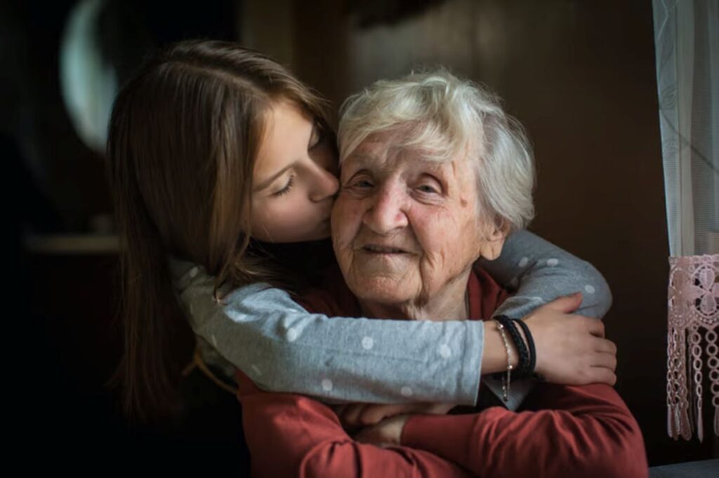 Young girl hugging and kissing her elderly grandmother on the cheek indoors, showing affection, love, and a close intergenerational bond.