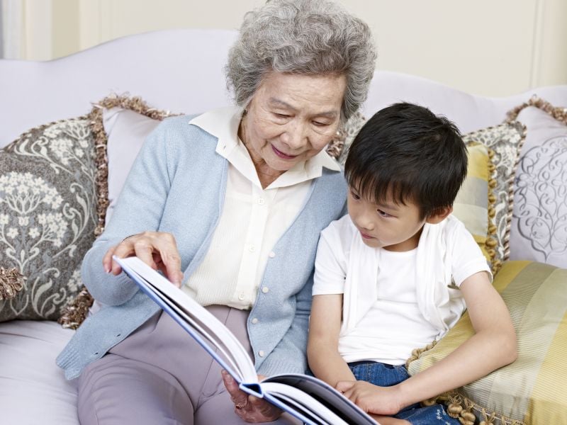 Grandmother and young boy sit closely on a sofa as she points to a page while reading a book together.