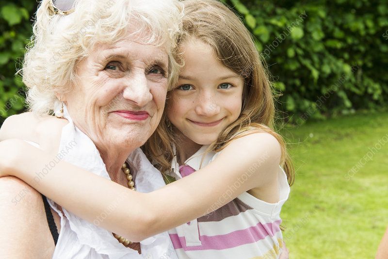 Young girl hugging her elderly grandmother outdoors, both looking at the camera with greenery in the background.