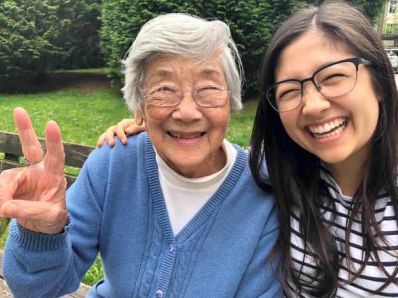 Grandmother and adult granddaughter smiling at the camera while sitting on a bench outdoors, with the grandmother holding up a peace sign.