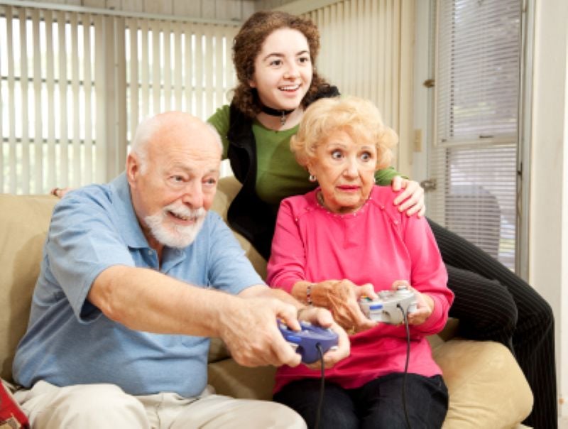 Psychologists Are Begging Grandparents To Do These 6 Things With Their Teen Grandchildren 3 Teen granddaughter standing behind her grandparents on a couch while they laugh and play a video game together in a cheerful family setting.