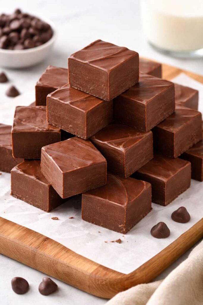 The Most Decadent 3-Ingredient Chocolate Fudge Recipe You’ll Ever Make 3 Neatly cut chocolate fudge cubes stacked on a wooden board with parchment paper and scattered chocolate chips nearby.