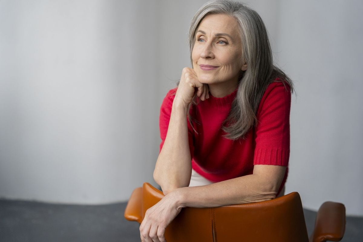 An older woman with long gray hair, wearing a red short-sleeve sweater, sits in a brown chair and rests her chin on her hand while gazing thoughtfully to the side against a plain background.