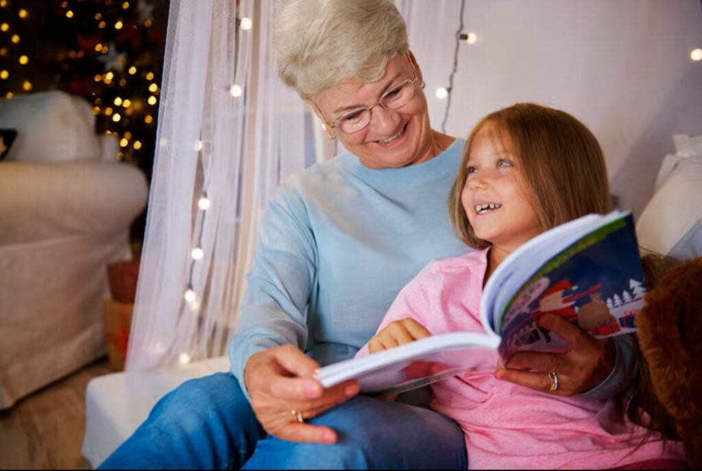 5 Small Things That Deeply Strengthen Your Bond With Grandkids, According to Psychology 4 Grandmother reading a colorful storybook with her granddaughter during a cozy evening indoors with warm lights in the background.