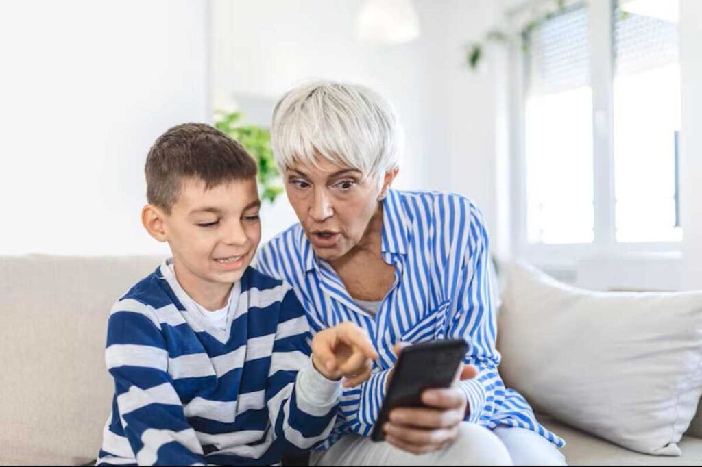 5 Small Things That Deeply Strengthen Your Bond With Grandkids, According to Psychology 3 Grandmother and young boy sitting together on a couch while looking at a smartphone, learning something together and sharing a curious moment.