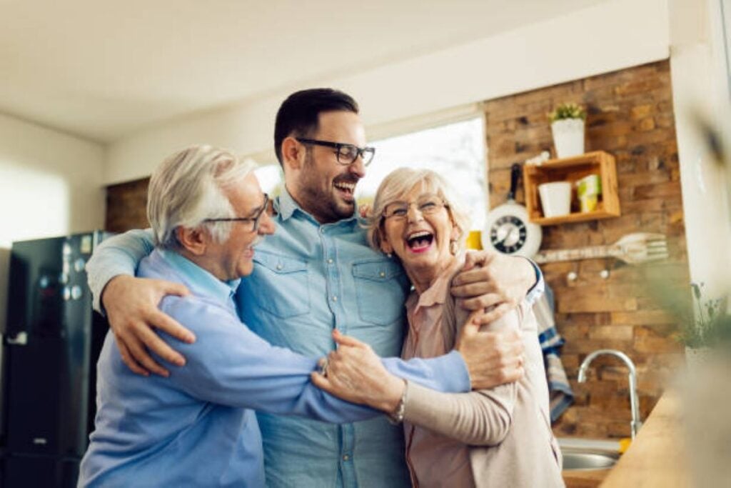 8 Things Older Parents Do That Make Adult Children Enjoy Visiting Them, According to Experts 3 An adult man laughing while hugging his elderly parents in a cozy kitchen, all three smiling and embracing each other affectionately.