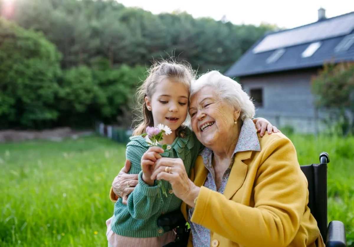 A smiling elderly woman sitting outdoors, embracing a young girl as they look at flowers together in a grassy yard with a house in the background.