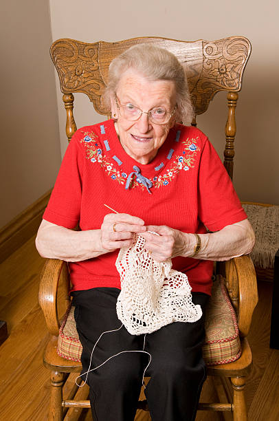 15 Addictive Grandmacore Hobbies That Feel Straight Out of a Simpler Time 4 Elderly woman sitting in a wooden chair indoors, knitting a lace piece with yarn and needles, wearing a red embroidered shirt and glasses.