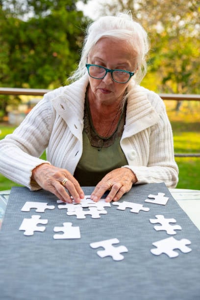 15 Addictive Grandmacore Hobbies That Feel Straight Out of a Simpler Time 3 Senior woman with white hair and glasses working on a jigsaw puzzle at an outdoor table.