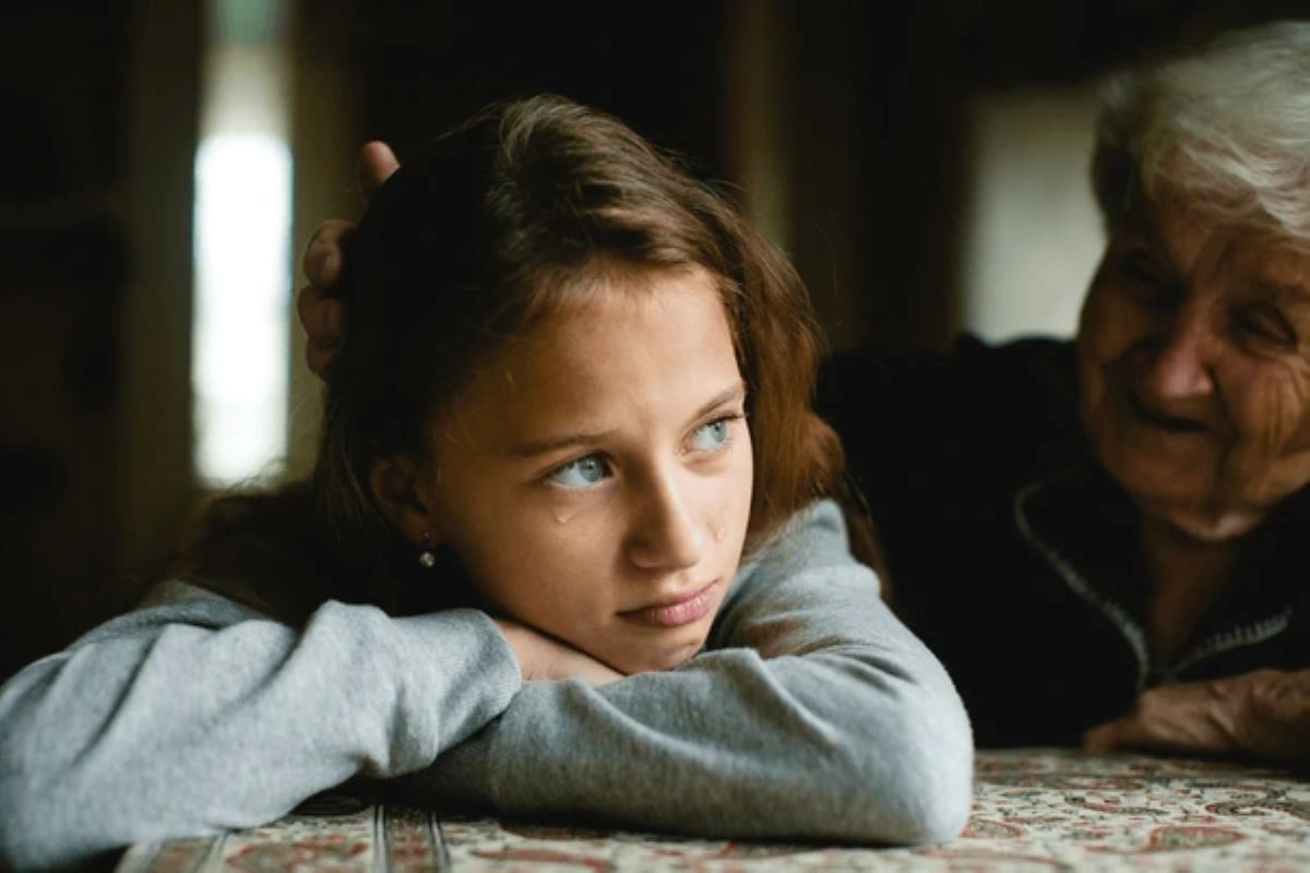 A young girl in a gray sweatshirt rests her head on her arms while an older woman beside her gently touches her hair and looks at her with concern.