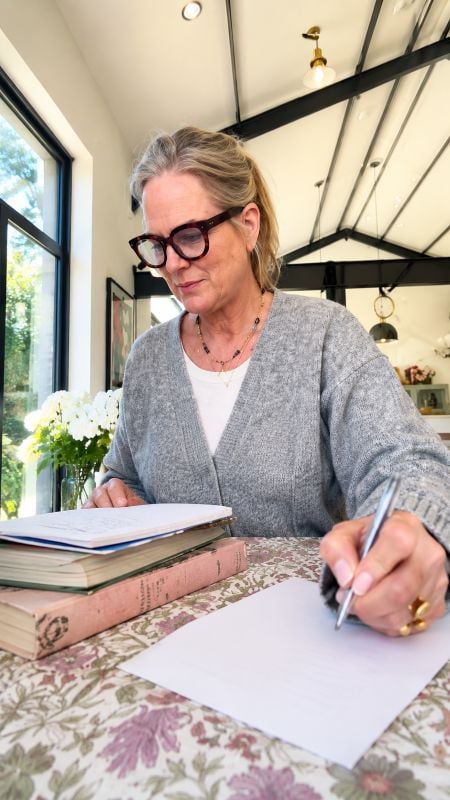 If you still write shopping lists on paper instead of using your phone, psychology says you have these 7 distinct qualities 4 A woman in glasses and a gray cardigan writes on blank paper at a floral-covered table beside stacked books, white flowers, and a bright sunlit window.