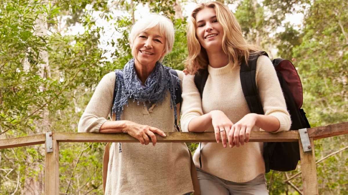 Older woman and teenage girl smiling together on a wooden bridge in a green wooded area.