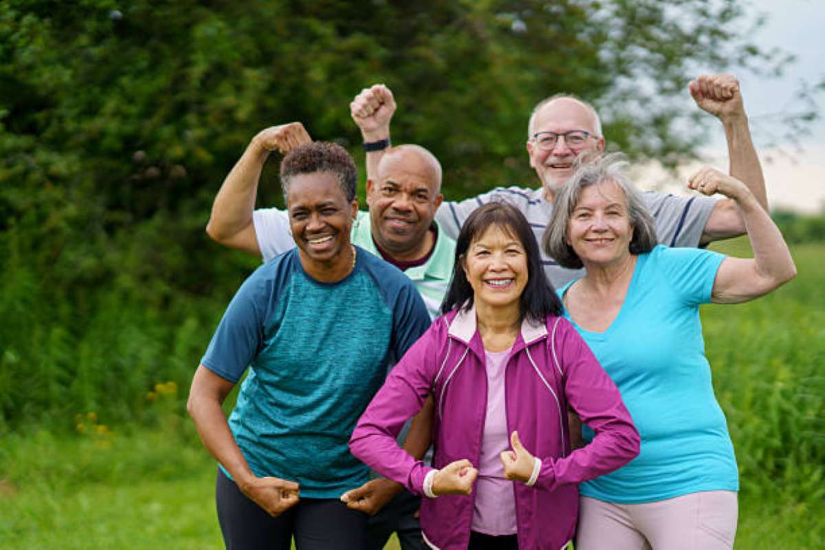 A diverse group of older adults smiling and flexing their arms together outdoors in a green park.