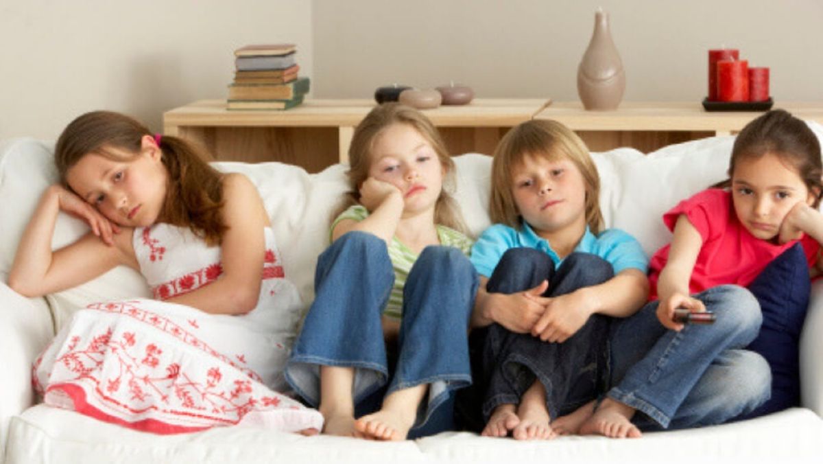 Four children sit slumped on a white couch, looking bored or tired, with books and candles on a shelf behind them.