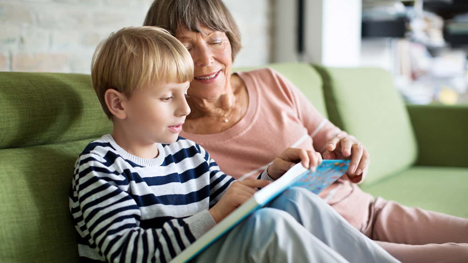 An older woman and a young child sit together on a green couch, looking at a picture book.