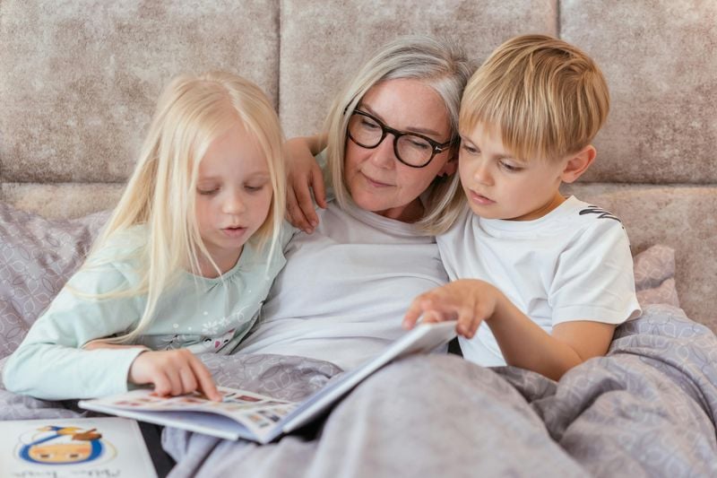 12 Things Grandkids Secretly Love Hearing From Their Grandparents, According to Psychologists 3 A gray-haired grandparent reads a picture book in bed with two young children.