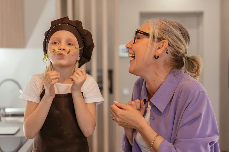 12 Things Grandkids Secretly Love Hearing From Their Grandparents, According to Psychologists 7 A laughing grandparent watches a child dressed as a chef play with a sprig of herbs in the kitchen.