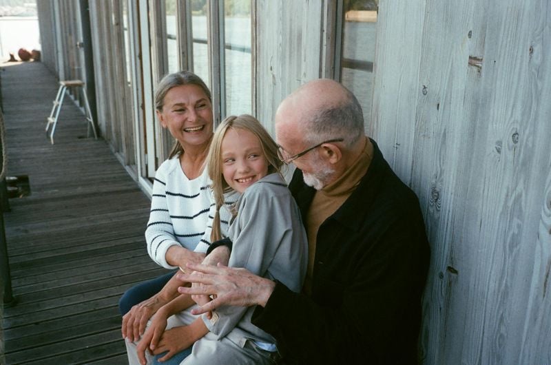 12 Things Grandkids Secretly Love Hearing From Their Grandparents, According to Psychologists 5 Two grandparents sit on a wooden porch with a smiling child between them.