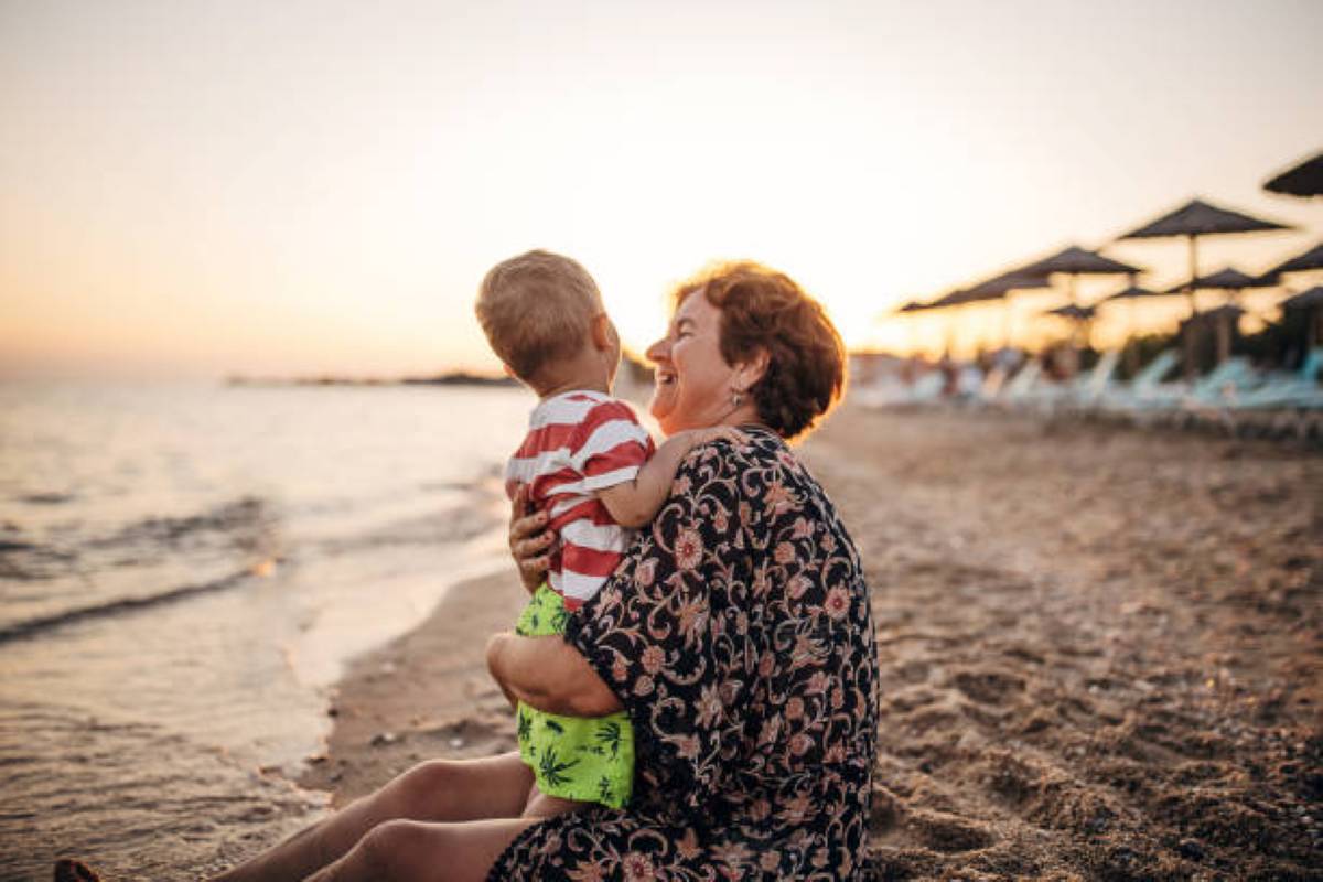 An older woman sits on a beach at sunset, laughing while holding a young child, with umbrellas and lounge chairs in the background.