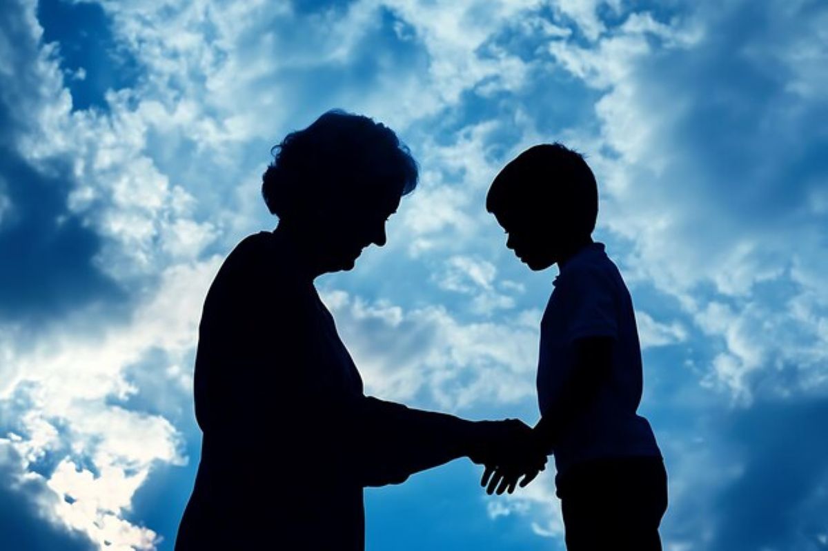 Silhouette of an older woman gently holding a young boy’s hand against a cloudy blue sky.