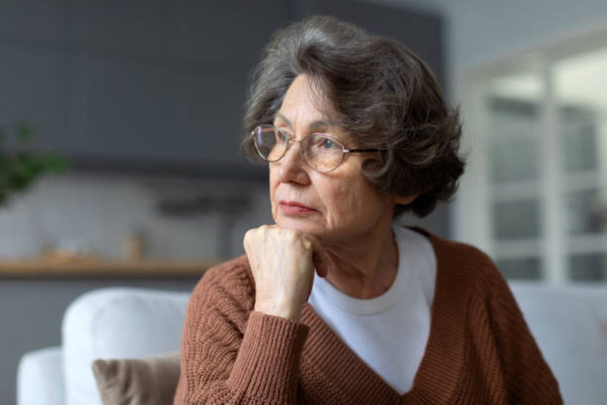 Older woman wearing glasses and a brown sweater rests her chin on her hand while looking thoughtfully out of frame in a softly lit living room.