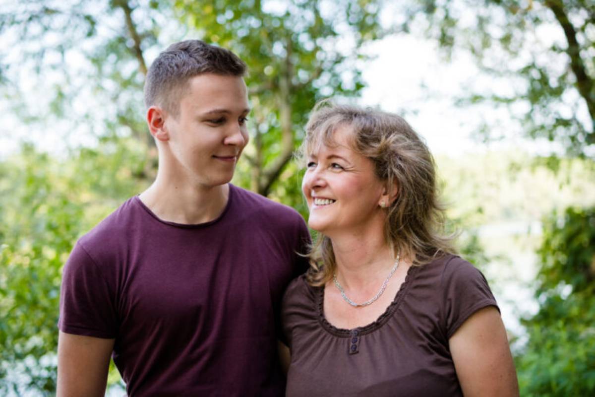 A young man and an older woman smiling warmly at each other while standing close together outdoors in a green, leafy setting.