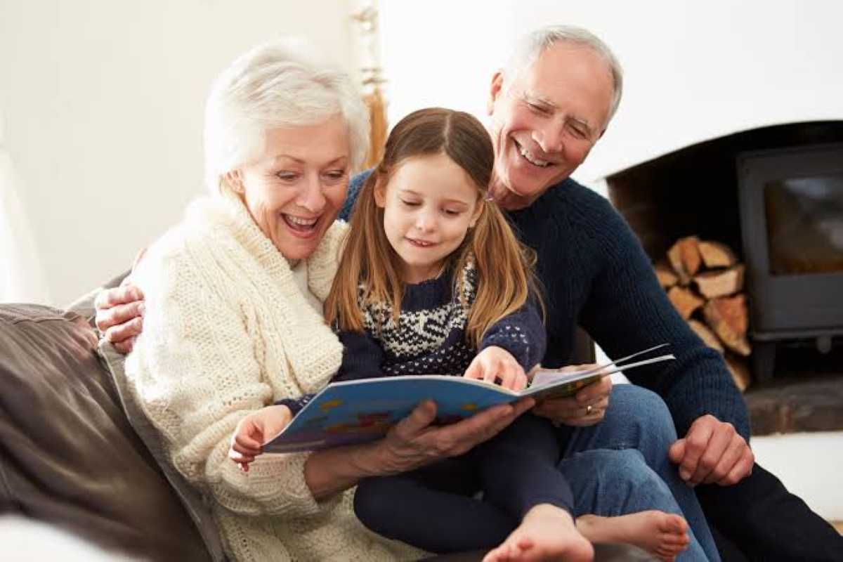 An older couple sits on a couch with a young girl between them, smiling as they read a picture book together near a fireplace.