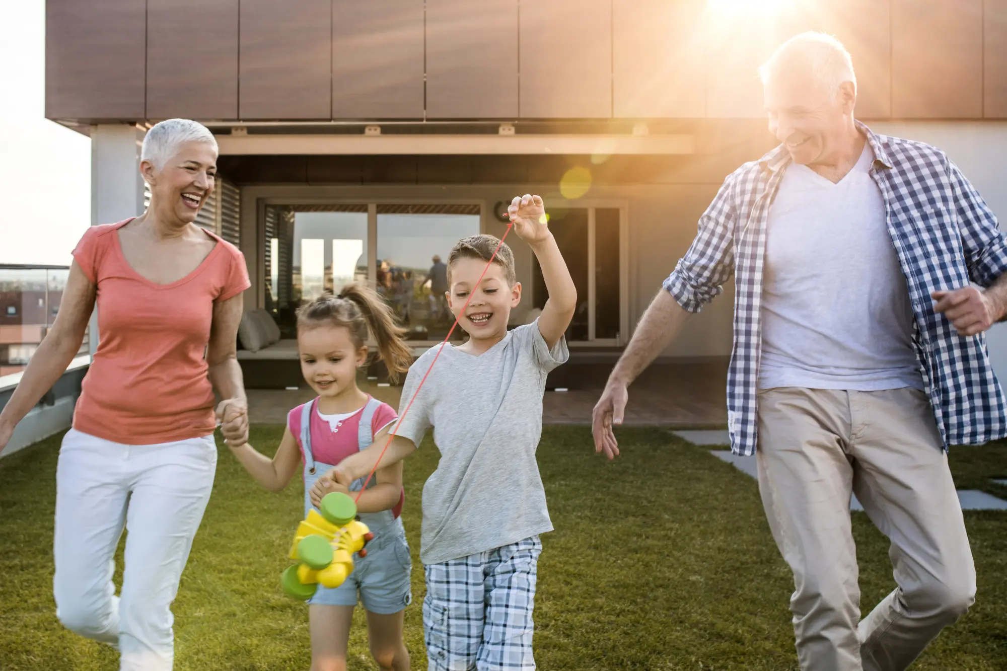 Older grandparents laughing and walking hand in hand with two young grandchildren on a sunny rooftop lawn, while one child pulls a toy on a string.