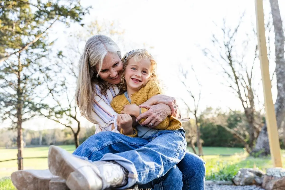 Grandmother hugging and smiling with her young granddaughter sitting on her lap outdoors in a sunny park, both looking happy and relaxed.