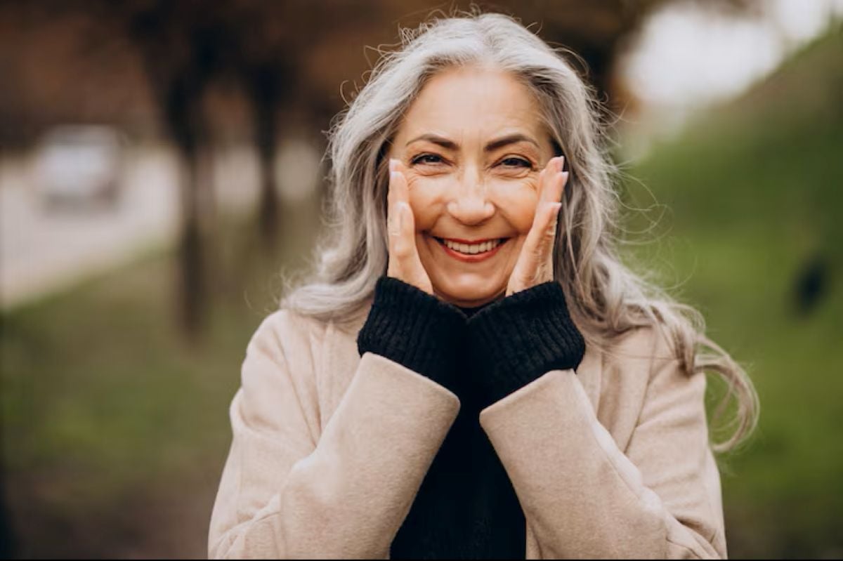 Smiling older woman with long gray hair, wearing a beige coat and black sweater, holding her hands to her cheeks outdoors with a blurred green background.