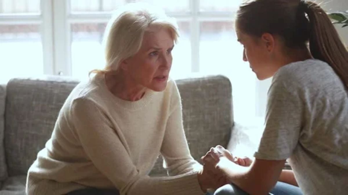 An older woman sits on a couch, holding both hands of a younger woman as they have a serious, supportive conversation in a softly lit living room.
