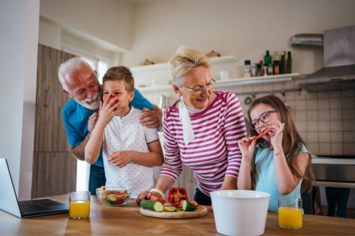 Grandparents and two children smiling together in a kitchen while preparing vegetables, with glasses of orange juice on the table.