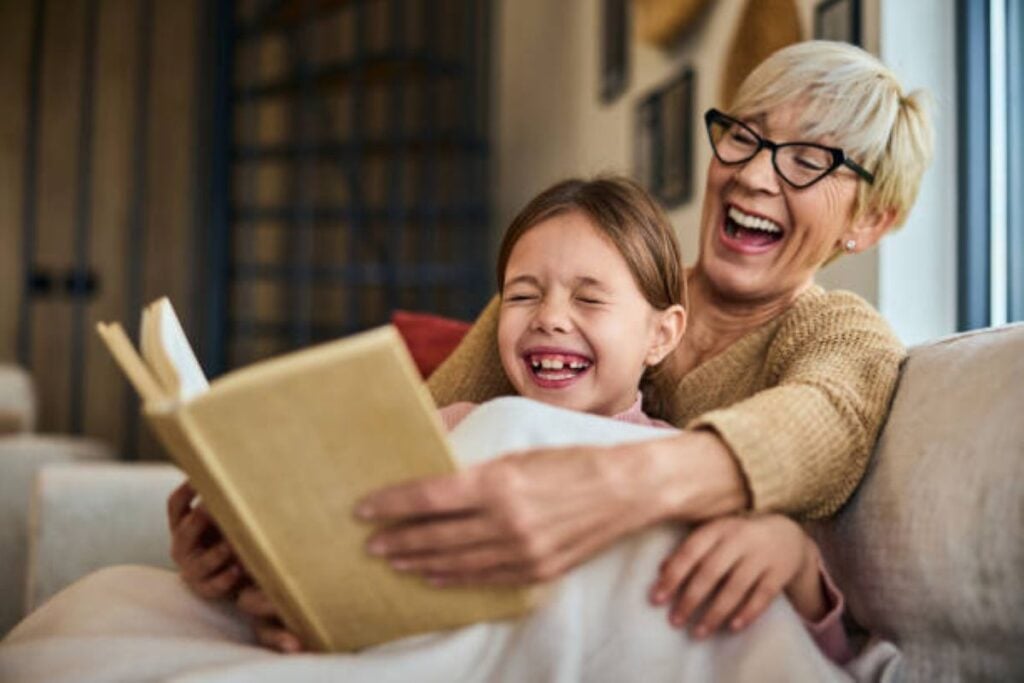 6 Secret Tricks for Grandmothers to Become Engaging Storytellers for Their Grandkids 4 Older woman with glasses laughing while reading a book with a young girl on a couch.