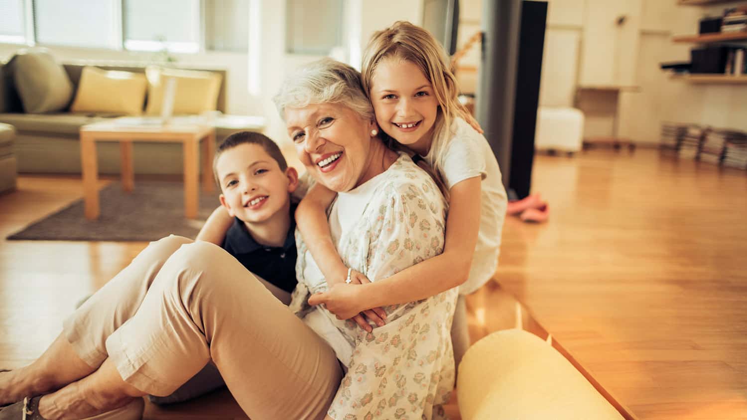 Older woman smiling while sitting on the floor and hugging two young children in a bright living room.