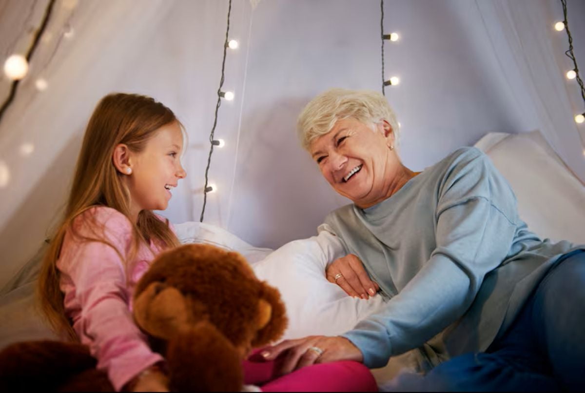 A grandmother and young granddaughter smile at each other inside a cozy blanket fort glowing with warm fairy lights, surrounded by soft pillows, a teddy bear, and bedtime comfort.