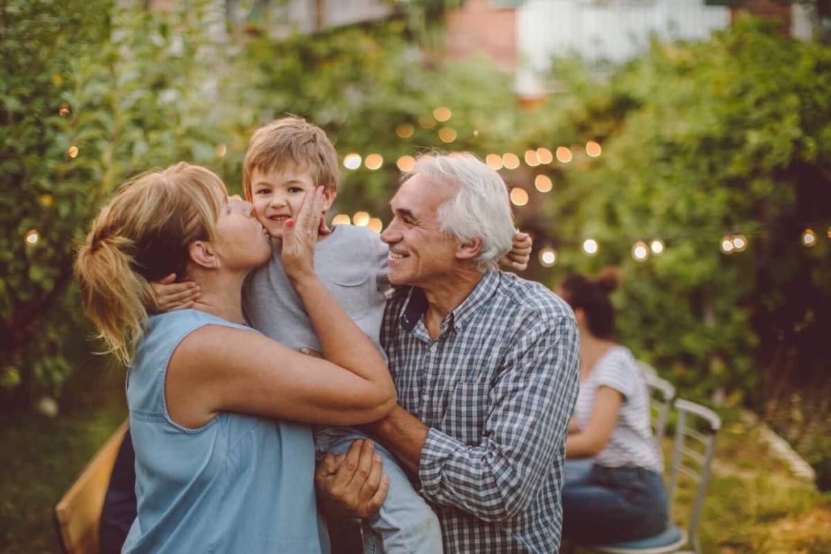 Woman kissing a young child on the cheek while an older man smiles beside them in a garden with string lights.