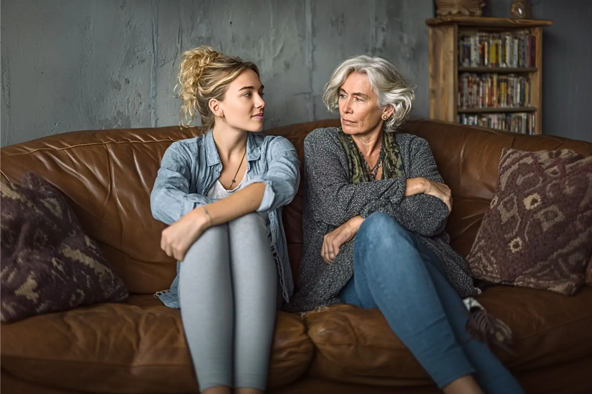 Two women sit on a brown leather couch in a living room, facing each other in conversation. The younger woman wears a light denim shirt and gray leggings, while the older woman has short gray hair and wears a gray cardigan with blue jeans. A bookshelf and patterned pillows are visible in the background.