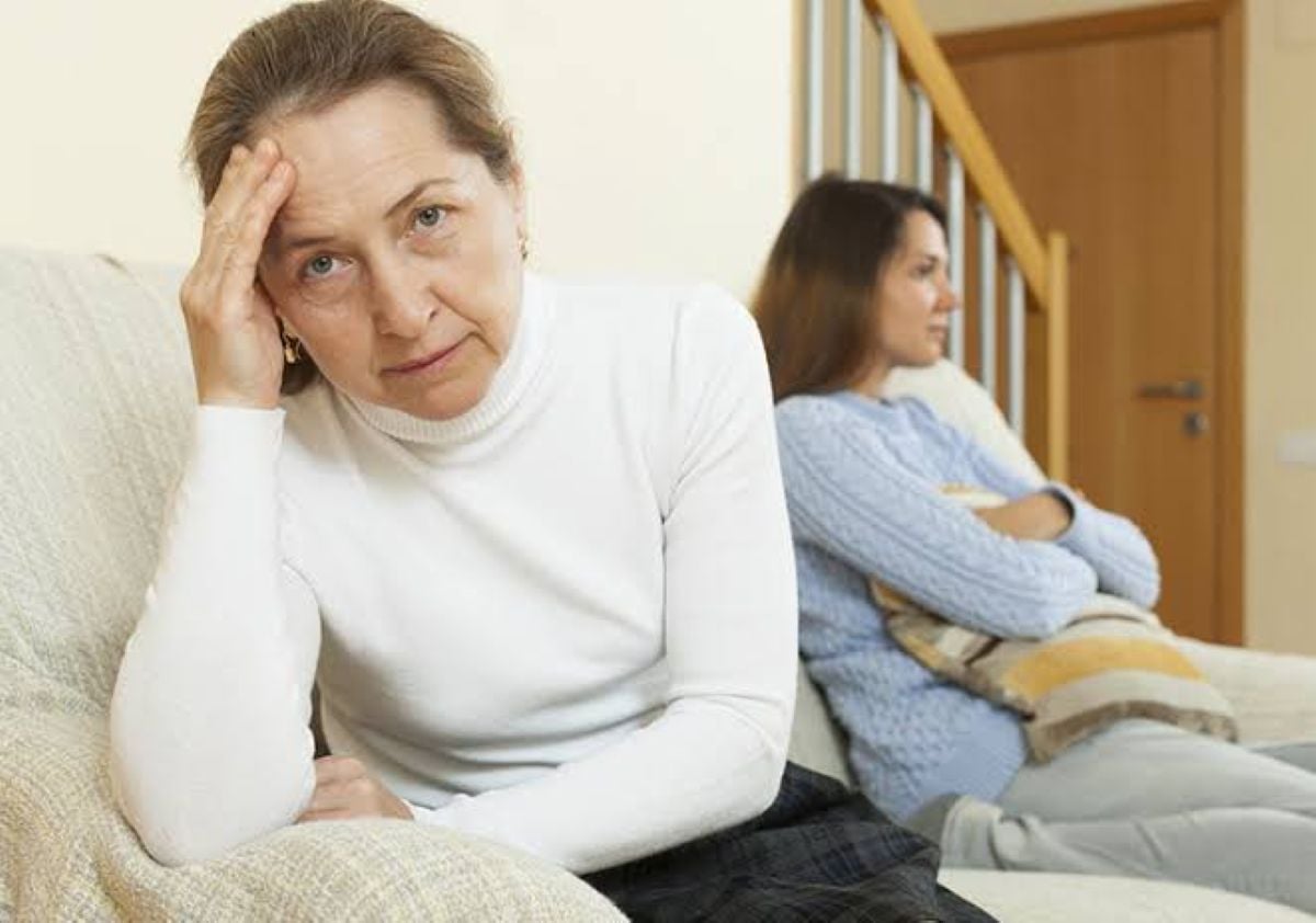 A worried older mother sits in the foreground with her hand on her forehead while her adult daughter sits turned away on a couch in the background, both appearing tense after a disagreement.