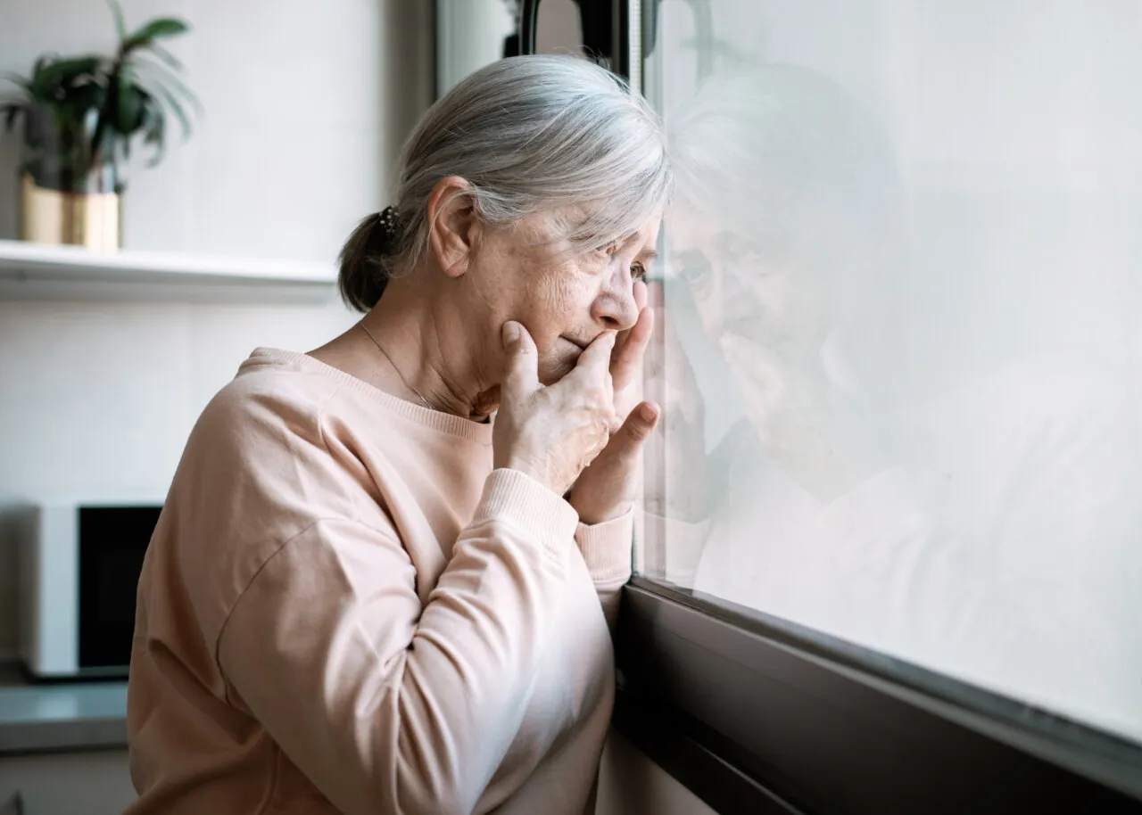 A senior woman standing by a window with her hand covering her mouth, looking outside with a sad, reflective expression.