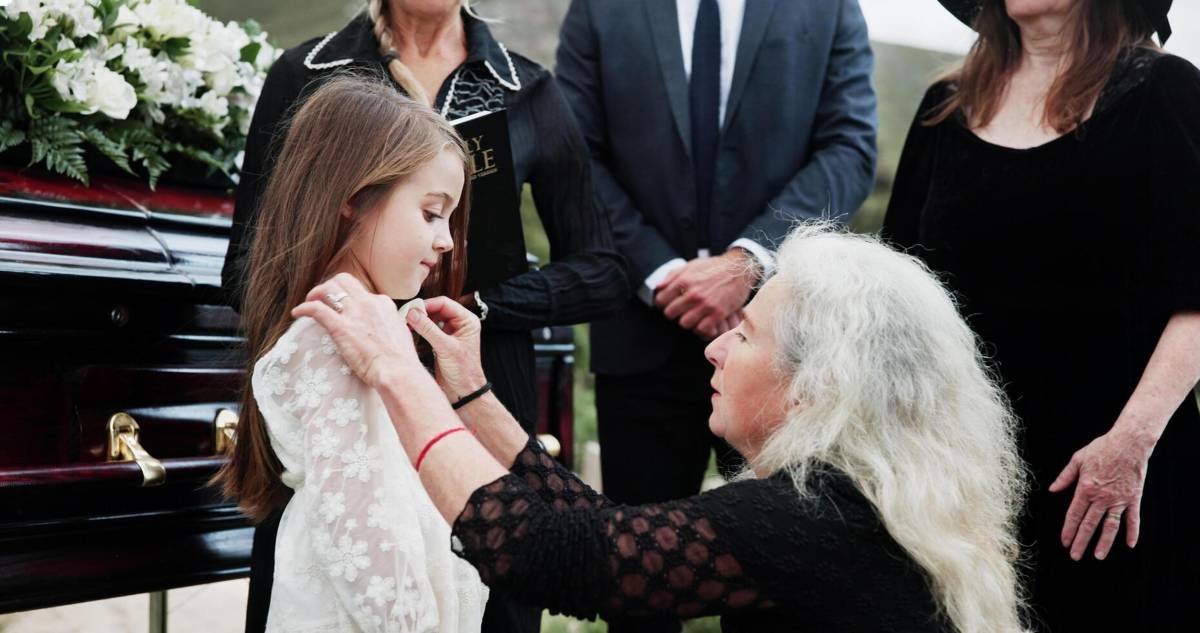 A grandmother gently adjusting a young girl’s dress beside a casket at a funeral, with family members standing nearby in a solemn setting.