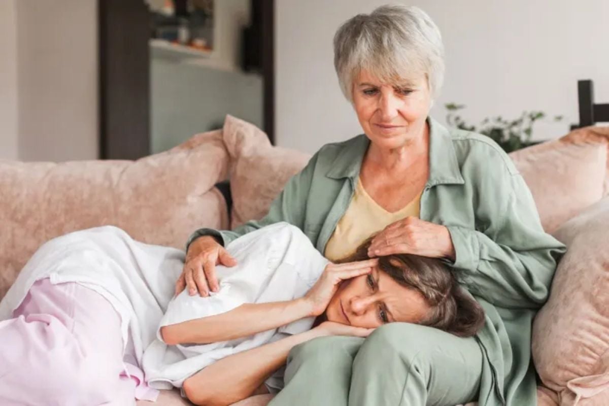 An older woman comforts a distressed younger woman resting her head in her lap as they sit together on a sofa.