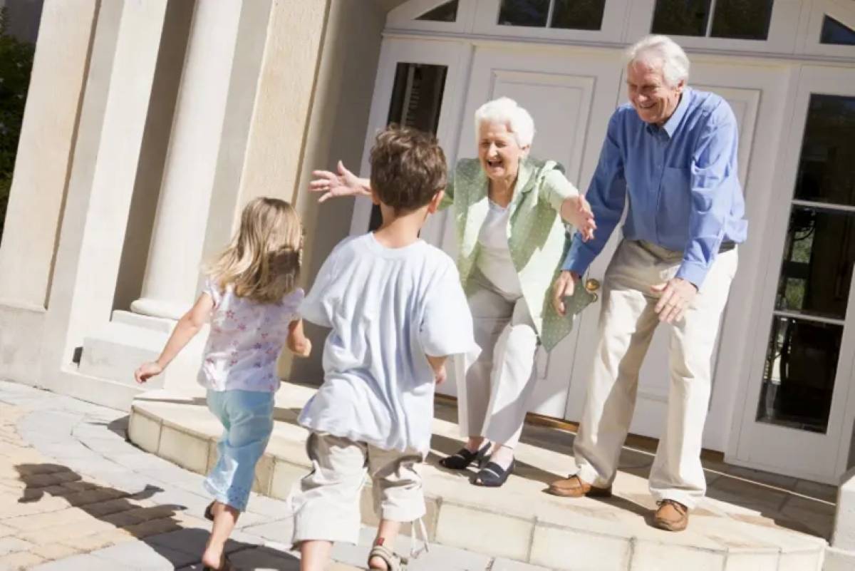 Grandmother and grandfather warmly welcome their two young grandchildren as they run toward them outside the front door of their home.