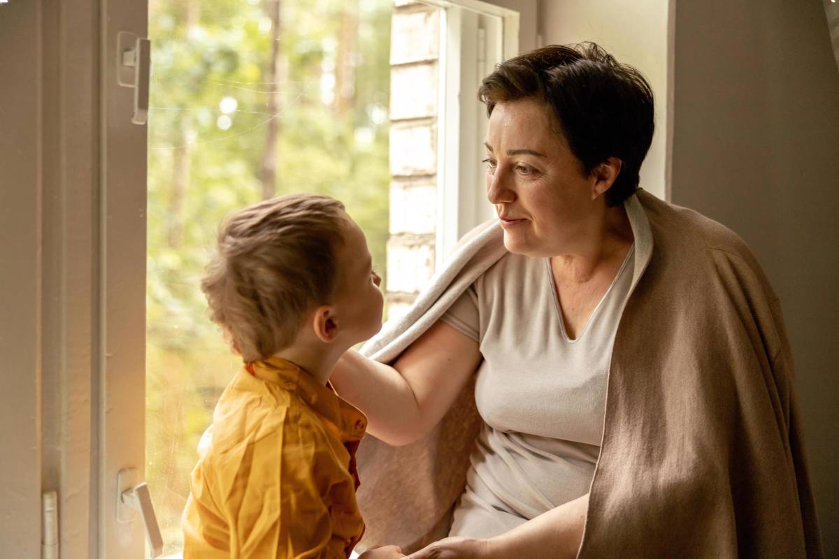 A grandmother and young grandson share a quiet, loving moment together beside an open window in warm natural light.