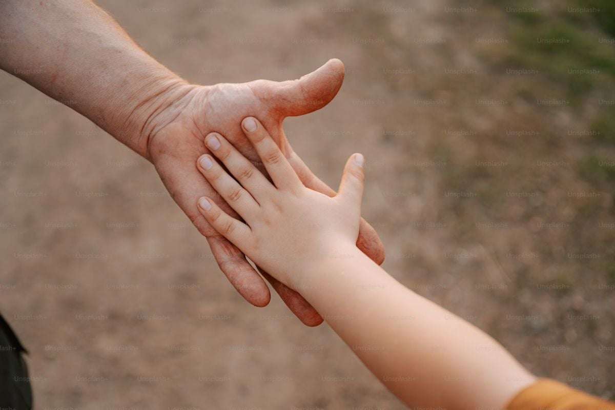 An adult gently holds a child’s hand outdoors against a softly blurred natural background.