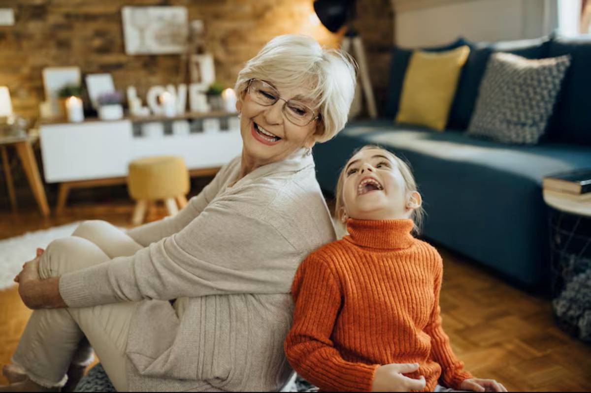 An older woman with short white hair and glasses sits back-to-back with a laughing young girl in an orange sweater, sharing a joyful moment in a cozy living room.