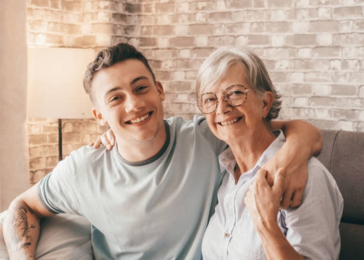 A smiling young man sits with his arm around an older woman wearing glasses as they pose together on a couch in a warmly lit room.