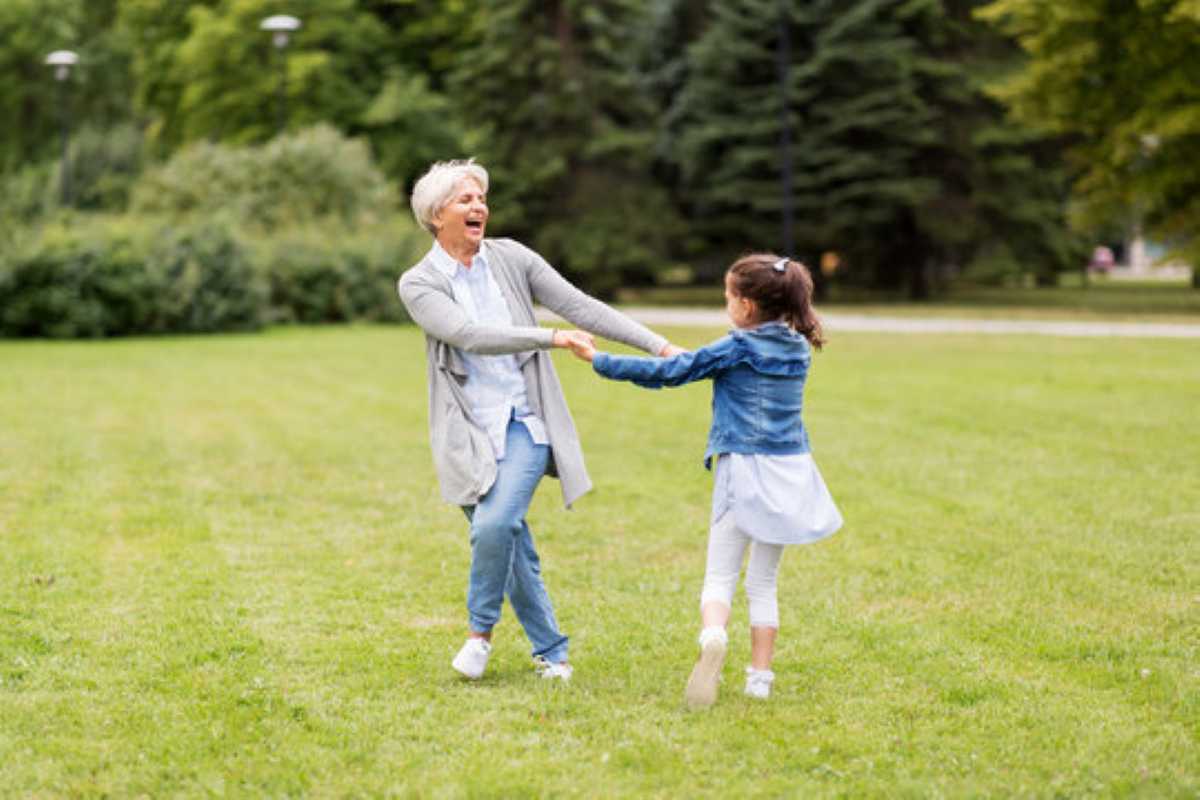 An elderly grandmother and her young granddaughter holding hands and joyfully spinning around together on a grassy lawn in a beautiful green park.