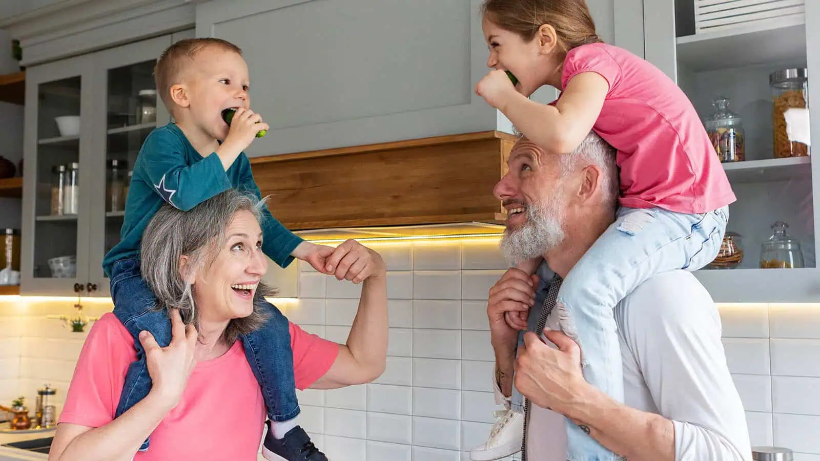 A smiling older woman and man carry two young children on their shoulders in a bright kitchen while the children eat cucumbers and the adults look up at them playfully.