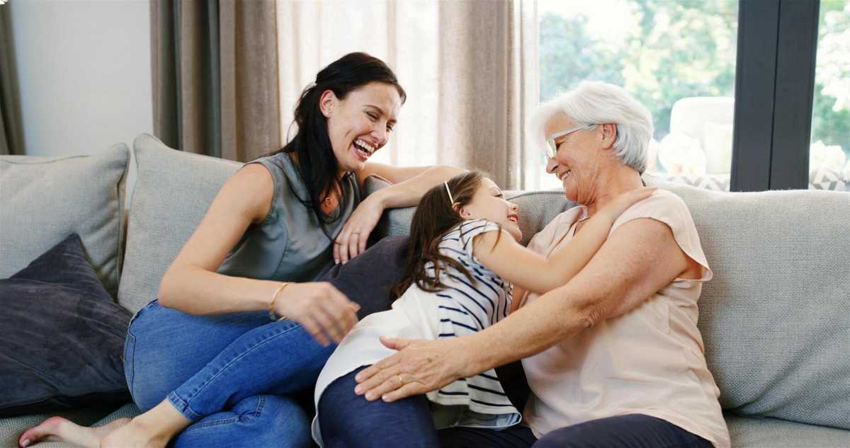Grandmother, mother, and young granddaughter laughing and cuddling together on a couch during a happy family moment.
