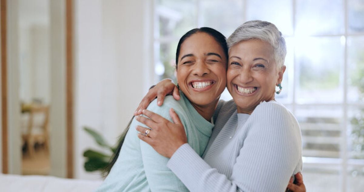 Two smiling women, a mother and her adult daughter, warmly embracing each other at home and showing a close, loving family bond.